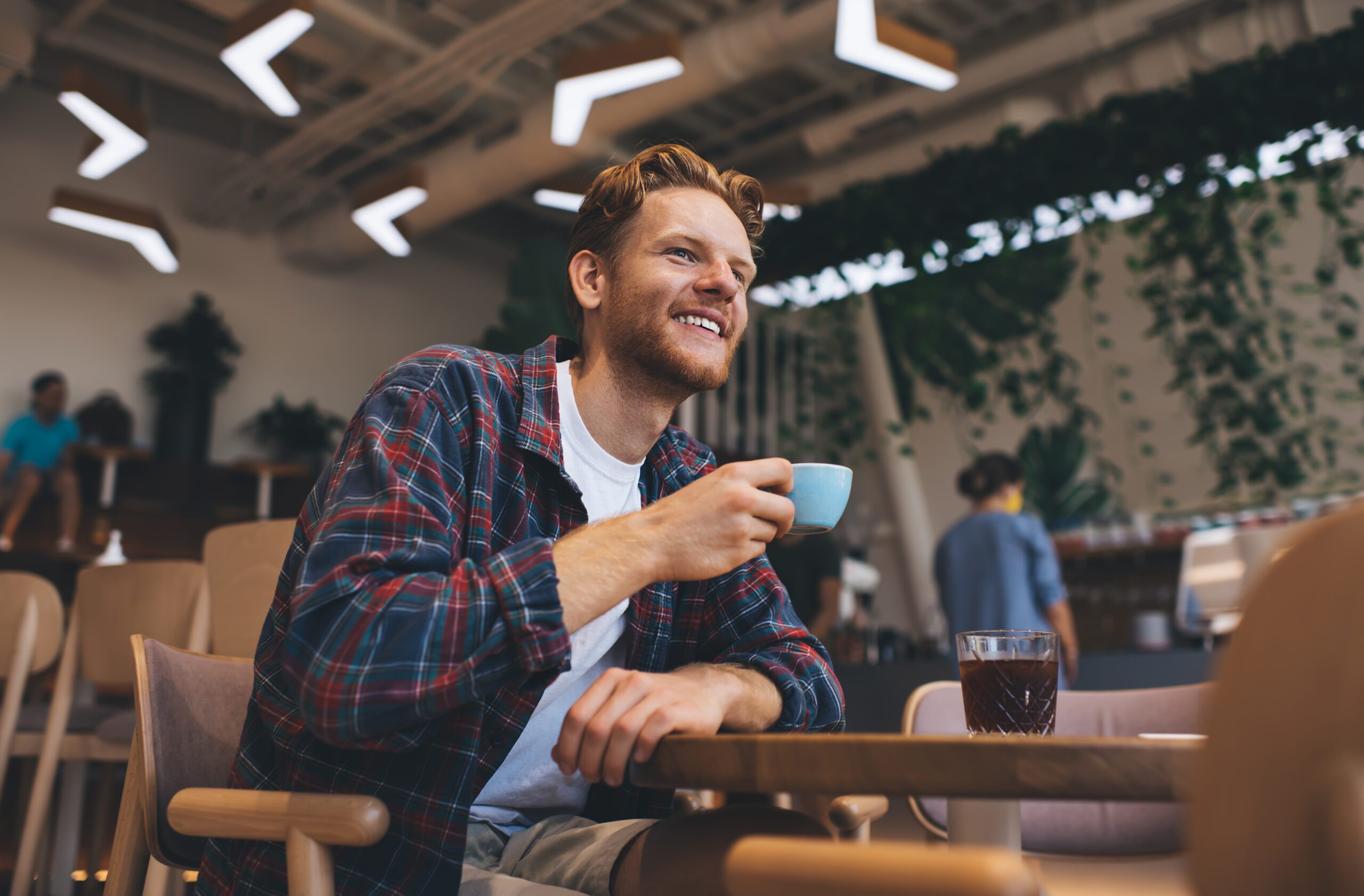 Young caucasian guy drinking tea or coffee from cup at wooden table in office space. Concept of rest and break at work. Idea of modern successful man. Smiling male person looking away