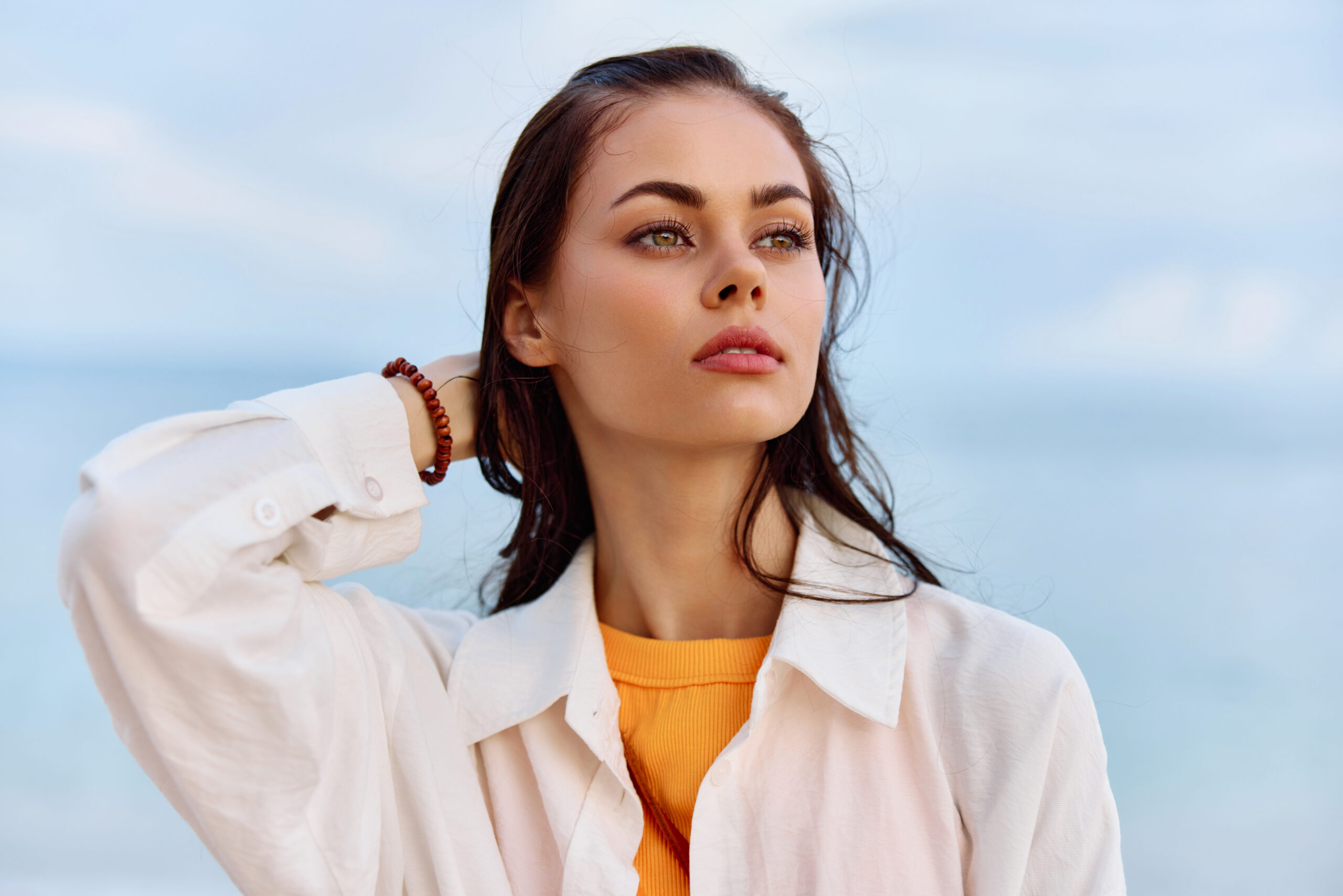 Portrait of a beautiful pensive woman with tanned skin in a white beach shirt with wet hair after swimming on the beach ocean sunset light with clouds. High quality photo