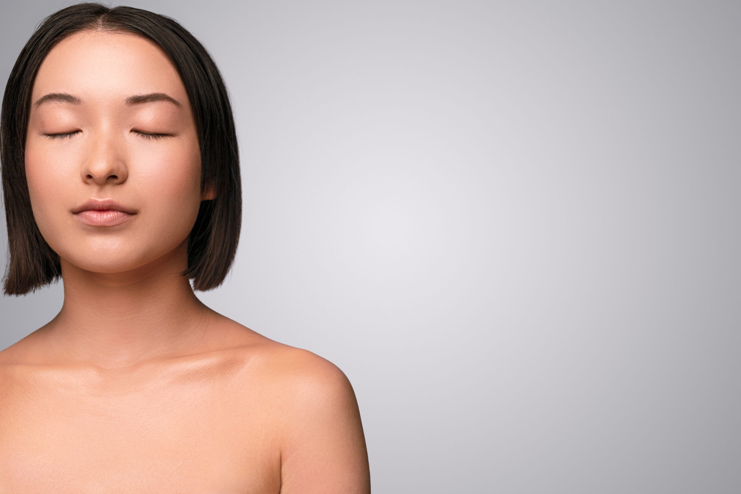 Young Asian lady with closed eyes and clean skin standing near blank space and representing spa industry against gray background